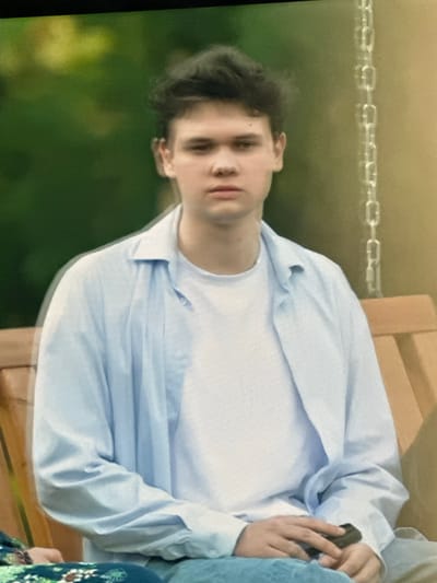 The image depicts a young person with dark, tousled hair sitting on an outdoor swing. They are wearing a light blue, unbuttoned collared shirt over a white t-shirt and blue pants. Their gaze is directed forward with a neutral expression. The background consists of blurred greenery and a chain, suggesting an outdoor setting possibly in a park or backyard. There is no visible indication of the city of Dondușeni, Moldova. The person is holding a dark object in their right hand.