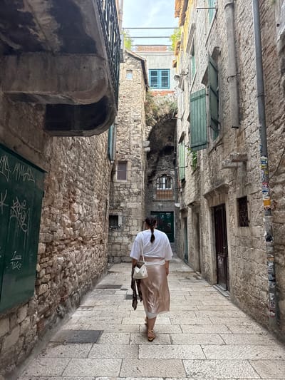 A woman with her back to the camera walks down a narrow, cobblestone alleyway in Split, Croatia. The alley is flanked by ancient stone buildings, some with green shutters and balconies. A large green sign with white graffiti is visible on the left wall. The woman is wearing a white top, a shimmering rose-gold skirt, and yellow sandals. She carries a white purse over her shoulder and a patterned scarf is draped around her neck. The sky is overcast.