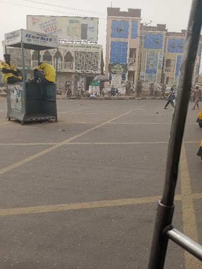 This image depicts an urban street scene in Kofar Mazugal, Nigeria, under an overcast sky. The foreground shows an asphalt road with prominent yellow painted lines, indicating traffic or parking demarcations. On the far right, a dark metallic structural component, likely part of a vehicle, partially obstructs the view.
In the midground, to the left, a grey, open-sided booth or stand labeled "Korbit" is visible. Two individuals, both wearing bright yellow long-sleeved shirts and dark trousers, are present at this structure. One is seated on the upper edge of the booth, facing right, while the second individual stands or leans towards the booth's interior, facing away from the camera. Advertisements for "Korbit Pure Water" are affixed to the booth's exterior.
The center midground features an open expanse of the asphalt road. A lone pedestrian in a blue shirt and dark trousers walks from left to right across this area. Further right, a partial view of a yellow vehicle and another pedestrian in a light-colored top are visible.
The background is dominated by several multi-story buildings. To the left, behind the "Korbit" stand, stands a building with a light-colored facade, characterized by arched window designs and intricate patterned screens on its upper levels. Above this building, a large billboard displays advertisements, including text that reads "NO MATTER THE MOOD... VIJU YOUR SIP" and the brand "BASTRI".
To the right of this, another prominent multi-story building features a brown-beige facade with numerous blue-paned windows and balconies. The names "KHADIJA" and "DAMKULLO" are visible on its upper sections. At street level in front of these buildings, there is considerable activity: several people are visible, some standing, others near what appear to be informal market stalls or setups, including one with a green umbrella. A person on a motorcycle is also present near these stalls. The overall scene suggests a busy commercial or public area.