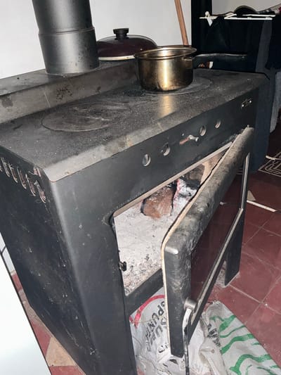 A black, wood-burning stove is positioned on a tiled floor in what appears to be a rustic kitchen in El Rodeo, Argentina. The stove has a visible chimney extending upwards and a partially open door revealing glowing embers and burnt wood within its firebox. A metal pot sits on the stovetop. The surrounding area shows signs of everyday use, with dust and ash on the stove and floor. Bags, possibly containing household goods, are placed near the stove.