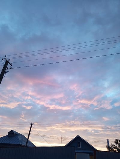 This is a photograph taken in Buraly, Russia, capturing a twilight sky filled with clouds tinged with pink and blue hues. In the foreground, a dark metal fence runs horizontally across the bottom of the frame. Silhouetted against the sky are the rooftops of two houses and a utility pole with power lines. The house on the left has a steep, metal roof with a dormer window. The house on the right is also characterized by a prominent gable roof, featuring a small, square window and a chimney. The overall scene depicts a rural or semi-rural residential area at dusk.