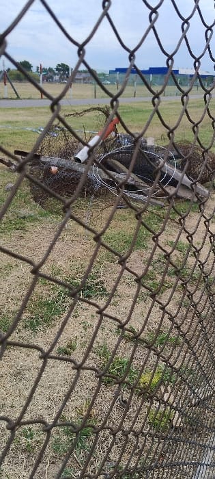 The image captures an outdoor scene viewed through a dark, weathered chain-link fence that occupies the foreground and extends diagonally across the frame. The fence's diamond-patterned mesh is in sharp focus, serving as a prominent visual barrier.

Beyond the fence, the ground is primarily covered with dry, yellowish-brown grass interspersed with patches of greener vegetation. In the mid-ground, several objects are discernible. To the left, a large, dark, seemingly discarded object, possibly a tire or rubber debris, lies on the ground. Wrapped around it and extending nearby is a coil of shiny, light-colored wire, which appears to be razor or barbed wire, suggesting a security feature. Adjacent to these items, a section of a white pipe with a distinct red-orange band near one end rests on the ground, along with a fallen wooden post or beam and other small debris.

Further in the distance, a long, low building with a prominent blue roof stands against the horizon. This structure suggests an industrial facility, a warehouse, or an aircraft hangar. A dark, possibly paved or gravel path runs horizontally across the mid-distance, separating the closer ground from the distant background elements. Beyond the building, a few trees are visible under a light blue sky, which shows minimal cloud cover.

The scene is illuminated by natural daylight, indicating either morning or afternoon. No people are visible, and no specific activity is taking place, presenting a static view of a boundary area. The presence of the security fence, the large open space, and the type of building are consistent with an industrial park or an airfield. Given the location context of San Fernando, Argentina, this setting strongly suggests an area around the San Fernando Airport (SADF). There is no visible text in the image.