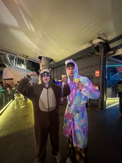 Two people pose in a vertically oriented photograph taken at night, likely in Grau i Platja, Spain. One person, wearing a hooded tie-dye robe and patterned pajama pants, stands slightly behind the other, who is dressed in a brown bear costume with sunglasses. The background shows what appears to be a street with buildings and lighting typical of an urban environment. The scene is somewhat brightly lit, suggesting an active outdoor space.