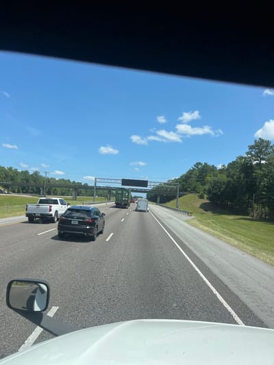 The image shows a view from the driver's seat of a vehicle on a highway, with the hood of the vehicle visible in the foreground. The road is asphalt with white lane markings. Several vehicles are traveling on the highway, including a white pickup truck, a black Lexus SUV, a green truck with a container, and a white trailer. Trees and vegetation line the side of the highway, and an overpass is visible in the distance. The sky is blue with scattered white clouds. The location is Riceboro, United States.