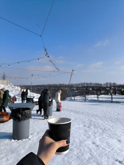 A forensic examination of the image depicts an outdoor, snow-covered recreational area in Prybuzke, Ukraine, under a clear, bright blue sky with minimal cloud cover.
In the immediate foreground, a fair-skinned right hand, adorned with dark blue nail polish, holds a black, corrugated disposable cup with a white interior and rim. A portion of a black jacket sleeve is visible at the bottom left.
The midground features a pristine expanse of snow. Several individuals, blurred due to depth of field, are present, dressed in winter attire. To the left, a person in a dark green coat walks past a gray cylindrical table topped with a black trash bag. Nearby, another identical table and trash receptacle are visible. Multiple blurred figures congregate around these tables. A person in a light-colored jacket stands with a child in a pink outfit adjacent to them, both facing away from the viewer. In the upper midground, strings of illuminated decorative lights are suspended between support poles, creating an overhead canopy.
The background reveals a distant treeline of bare, dark trees stretching horizontally across the horizon. Below this, a gentle slope of snow leads to a blurred, distant landscape. To the right, a low, light-colored wooden fence runs parallel to the horizon. Additional blurred figures are visible in the far background, possibly engaging in activities on the snowy terrain. The overall scene suggests a sunny winter day at a public or recreational facility.