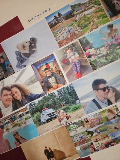 The image displays a wall-mounted collage of approximately 20-25 personal photographs, arranged diagonally and overlapping, on a light-colored surface with a visible deep red section on the left and bottom. The word "Memories" is spelled out near the top center using individual letter cutouts. This display is located in Cherwell District, United Kingdom.
The collage content depicts diverse scenes:
* A person playfully interacting with a golden retriever in deep snow.
* A couple walking on a path overlooking a coastal landscape.
* A picturesque townscape with white buildings.
* A woman sitting on a rock overlooking a scenic vista.
* A woman in a swimming pool, viewed from behind, with pink and white orchids in the foreground.
* A man in glasses and a Santa hat smiling in a festive indoor setting.
* A child in a Santa hat and patterned pajamas holding an object in front of a Christmas tree.
* A young man and woman embracing and smiling.
* A white vintage Volkswagen camper van parked in a grassy area, with a woman holding a golden retriever next to it.
* A man in sunglasses kissing a woman on the cheek on a beach.
* A man and woman in a swimming pool.
* Two individuals standing against a textured wall, casting long shadows.
* Smaller photos include a woman in a field of purple flowers, mountainous landscapes, a couple walking over a bridge, coastal views, and other casual outdoor and indoor gatherings.