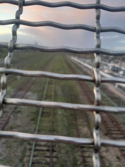 An eye-level, close-up shot captures a section of a metal grid fence. The focus is sharpest on the fence, while the background is blurred. Parallel train tracks stretch into the distance, bordered by vegetation. A building is faintly visible in the top center. Soft, muted colors dominate the background, suggesting either a sunrise or sunset. The image has a slightly disorienting, tilted perspective.