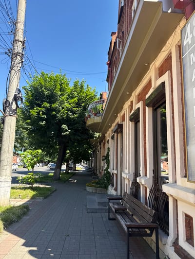 The image depicts a street scene in Sunzha, Russia, on a clear day with a bright blue sky. In the foreground, a grey concrete utility pole with numerous black electrical wires is prominent on the left side. Adjacent to the pole is a sidewalk paved with grey rectangular bricks. A dark wooden bench with a metal frame is situated on the sidewalk next to a building. The building itself has a facade of red brick and decorative white columns around its windows. Several windows on the building are fitted with dark brown roller shutters. A large green tree with lush foliage is located on the left side of the sidewalk, casting shadows on the pavement. In the background, a street with cars and other buildings can be seen. The overall impression is a typical urban streetscape.