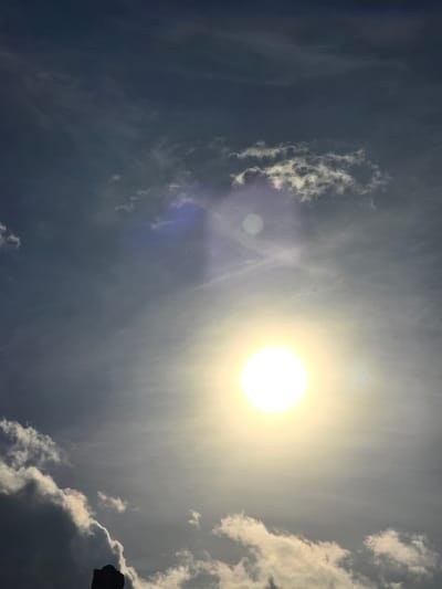 This media file is a low-angle, wide shot of a bright, hazy sun in a cloudy sky. The sun is the dominant subject, appearing as a glowing, yellowish-white orb in the upper right quadrant of the frame. Thin, wispy clouds are scattered across the sky, some illuminated by the sun, while others appear darker and more dense, particularly on the right side. A faint lens flare is visible, creating a circular, purplish-blue smudge in the lower left portion of the image, and another less distinct halo of light. The overall atmosphere is bright and somewhat diffused due to the haze.

The scene depicts an outdoor setting, likely during the daytime, given the presence of the sun. The time of day appears to be mid-day or late afternoon, judging by the brightness and position of the sun. The weather is partly cloudy. There are no people or specific objects clearly identifiable in the frame, except for a dark, silhouetted shape of what appears to be the edge of a structure or natural formation on the far right edge of the image, extending slightly into the frame. The image evokes a sense of warmth and natural light. No text is visible in the media file.

The provided location context places this scene in Kostelec na Hané, Czechia.