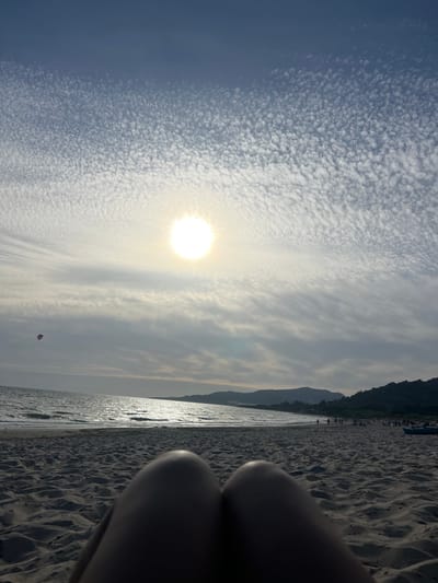The image depicts a view from the beach in Tarifa, Spain. The foreground is dominated by the textured sand and the blurred legs of the photographer. Beyond the sand, the sea stretches out, with gentle waves reflecting the bright, slightly hazy sun positioned high in the sky. The sun is surrounded by wispy clouds that form intricate patterns across the blue sky. In the distance, a line of hills and mountains forms the coastline. The beach appears to be populated by several figures, some walking along the shore and others gathered in groups. A kite is visible in the air to the left of the frame. A blue boat is present on the sand to the right.