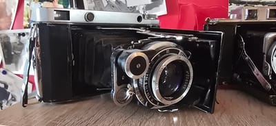 The image displays two vintage cameras on a wooden surface. The camera in the foreground is a black bellows camera, open and partially extended. It features a silver lens with various markings including aperture and distance scales, as well as a serial number that appears to be "N° 208046". A small winding knob is visible to the left of the lens. The top of this camera has a silver rectangular element with two circular indents. The second camera, to the right, is also black with silver accents and appears to be a box camera with a viewfinder. In the background, unfocused elements suggest a collection of old photographs and a bright red object, possibly fabric or paper. The scene is lit from the front, casting soft shadows. The location of Gostagayevskaya, Russia, is not determinable from the visual information provided in the image.