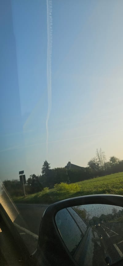 The image, likely taken from inside a moving vehicle, shows a landscape through the passenger side window. The sky, a gradient from a light blue to a hazy off-white, dominates the left side, with a thin contrail bisecting the area horizontally. The right side features green foliage, likely trees and fields, running along a road. A car side mirror occupies the extreme right of the frame. The reflection within the mirror shows a blurred scene of the same landscape, possibly with other vehicles. The image is rotated 90 degrees counter-clockwise. The location is speculated to be in the city of Broadland, United Kingdom, although no specific identifiable landmarks are visible.