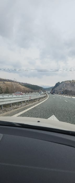 The image depicts a multi-lane asphalt highway viewed from the interior of a vehicle, specifically from its dark grey dashboard and lower windshield. The scene is located on a highway near Staro Selo, Bulgaria, under an overcast sky with light grey clouds.
The highway stretches into the distance, showing signs of a traffic slowdown or congestion with multiple vehicles, including passenger cars and at least one commercial truck, visible in the far lanes. A red passenger car is parked or stopped on the left shoulder or median area.
To the left of the roadway, a metal crash barrier runs parallel to the direction of travel, separating the highway from a sloping hillside covered with bare deciduous trees and scattered evergreens. Patches of snow are visible on the ground. To the right, another sloping hillside features sparse vegetation, exposed earth, and additional snow patches. A blue road sign is partially visible on the right side of the highway further down the road. In the far background, snow-capped mountains are discernible beneath the cloudy sky. No identifiable individuals are visible in the scene.