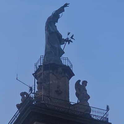 The image shows a statue of a religious figure, likely Jesus Christ or a saint, standing atop a column or tower. The figure is cloaked and holding a branch with leaves in its left hand, while its right arm is raised. A halo is visible around its head. The statue is made of a dark material, possibly bronze or stone.

At the base of the main statue, there are other sculptures of angelic figures. One is on the right, appearing to be a winged cherub or angel sitting down. Another figure, on the left and lower down, is also seated and appears to be a cherub or putto, with its back to the viewer.

The statues are situated on a structure with a decorative metal railing. The background is a clear, pale blue sky, suggesting it is daytime or early evening. There are no people visible in the image. The architecture appears to be of classical or religious style. The image does not provide information about the specific location in Calcinate, Italy, only that it depicts a statue or monument.