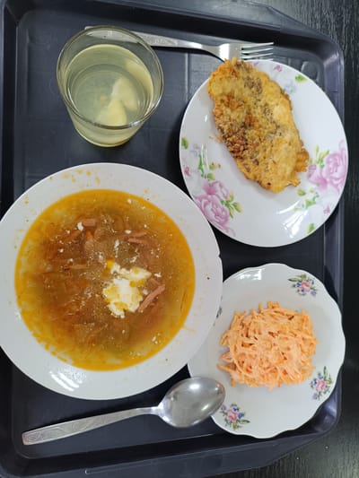 A top-down, medium shot captures a meal served on a black tray. On the tray, from left to right, are a glass of pale yellow liquid with ice, a large white bowl filled with a golden-orange soup containing visible meat and a dollop of white cream, a small white plate with a breaded piece of food, and a smaller white bowl filled with shredded orange salad. A silver fork rests above the breaded food, and a silver spoon lies to the left of the soup bowl. The tray is positioned on a dark surface, possibly a table. This scene is set in a cafeteria or dining hall, likely in the city of Dnestrovsk, Moldova. The lighting is even, and there are no people visible in the frame.