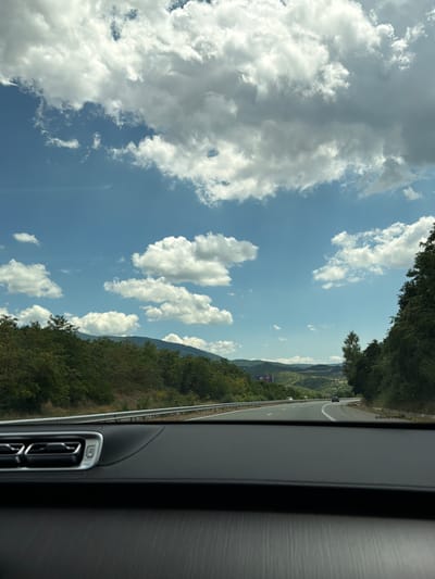 The image shows a view from inside a car driving on a highway. The highway curves to the right, with a metal guardrail on the left. Lush green trees and rolling hills covered in vegetation line both sides of the road, with mountains visible in the distance. The sky is a bright blue with scattered white clouds. The foreground of the image is dominated by the car's dashboard, which is black and has some metallic accents. There is no visible indication of people in the image. The scene appears to be set in a rural area outside of Djerman, Bulgaria, based on the provided location context.