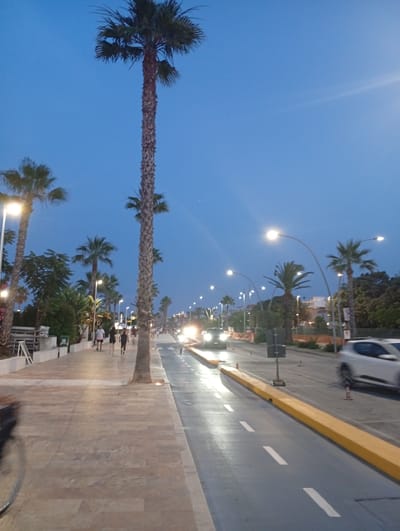 The image depicts a street scene at dusk in Civitanova Marche, Italy, likely along the Lungomare, a promenade area. A wide, paved pedestrian walkway runs along the left side of the image, lined with tall palm trees and streetlights. Several people are walking on the pedestrian path, appearing to be enjoying a stroll. Across from the walkway is a road with vehicles, including a white car in motion on the right side, and traffic lights. The road is separated from the walkway by a raised yellow barrier. The sky is a clear, deep blue, indicating the transition from day to night. The overall atmosphere is calm and leisurely, suggesting a typical evening in a coastal Italian town.