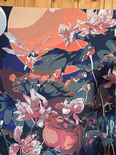A partially completed paint-by-numbers canvas depicting a natural pond scene is presented, observed in 3-Y Uchastok, Russia. The canvas rests against a light-colored wooden surface, visible at the top edge of the image. The artwork features a prominent reddish-brown frog in the lower-right quadrant, its body mostly painted, facing right. Multiple pink and white water lilies, some fully painted and others displaying numbered sections (e.g., 15, 20), surround the frog and are scattered across the lower and central portions of the canvas. In the upper-left quadrant, a light blue and grey dragonfly with segmented wings is depicted in flight, with several numbered areas (e.g., 16, 17, 36) still unpainted on its body and wings. The background includes a large, circular, coral-orange shape, suggestive of a celestial body like a sun or moon, with a partial white circle visible in the extreme upper-left corner. The overall environment is characterized by dark blue, dark green, and purple tones representing water and foliage. The canvas displays visible black outlines and numerical indicators for unpainted regions across the entire scene.