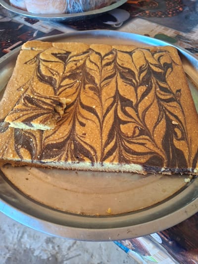 A marbled cake with a slice removed is presented on a round metal platter. The cake exhibits a swirl pattern of light brown and dark brown batter. The platter is set on a patterned surface. The background suggests an indoor setting with various objects and textures. The scene does not depict any specific location within Ko`kdala, Uzbekistan, or any individuals.