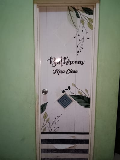 The image shows a white interior door with a wooden frame, set against a light green wall. The door is decorated with dark green leaf and vine decals, and the words "Bathroom" and "Keep Clean" are written in a black, cursive font. A silver doorknob is visible on the left side of the door, slightly below the center. There is a square, diamond-patterned decal in blue and white on the lower half of the door. The bottom of the door has a decorative stripe pattern of black and gray, with some areas of peeling. The scene is indoors, and appears to be a typical residential setting in Sendangrejo, Indonesia. There are no people visible in the image.