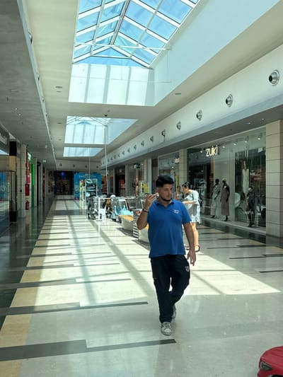 A man in a blue polo shirt and black pants is walking down a brightly lit shopping mall corridor. The mall has a high ceiling with recessed lights and large windows on one side that let in natural light. The floor is made of light-colored tiles with dark stripes, creating a patterned effect. The man is holding a small white object in his right hand near his mouth and appears to be looking at it.
In the background, several shop windows are visible, displaying clothing and mannequins. One shop sign reads "ZUKI." Other signs and posters are partially visible further down the corridor, with some text in Italian. Other people can be seen in the distance, shopping or walking. The scene suggests it is daytime, with bright light casting shadows on the floor. The overall atmosphere appears to be casual and ordinary, as one might expect in a shopping center. The man's expression is neutral.