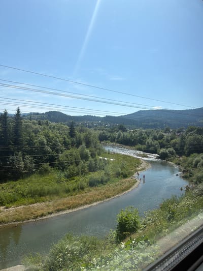The image shows a wide, winding river flowing through a valley with lush green trees on either side. In the foreground, the river is calm and reflects the clear blue sky. There are several power lines stretching across the frame. In the middle ground, a few people are standing in the shallow water. Beyond the river and trees, a village with houses is visible nestled in the foothills of mountains. The overall scene is one of natural beauty and a peaceful rural setting. The location is identified as the city of Mykulychyn, Ukraine, which is known for its scenic Carpathian Mountain landscape.