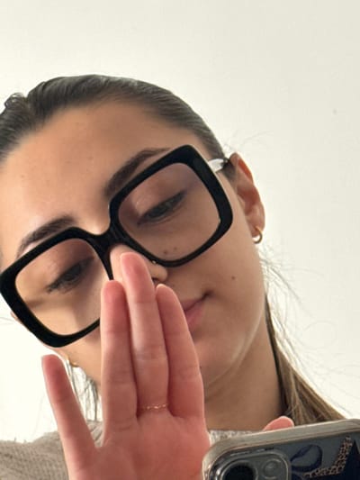 The image displays a close-up, low-angle shot of an adult female, positioned in an indoor setting in Limeil-Brévannes, France. The subject wears large, square-framed black eyeglasses. Her dark hair is pulled back, and small gold-colored hoop earrings are visible on both ears. Her gaze is directed downwards. A faint smile is perceptible on her lips. Her right hand is raised, palm partially facing the camera, covering the lower portion of her face from the nose down. A thin, gold-colored ring with a subtle textured design is present on the ring finger of her right hand. The lower right corner of the frame reveals the top portion of a smartphone, held by the subject's left hand. The phone is encased in a transparent cover revealing a leopard print pattern beneath, and its multi-lens camera module is visible. The subject wears a light-colored, possibly knitted, garment. The background is a plain, undifferentiated light-colored surface.