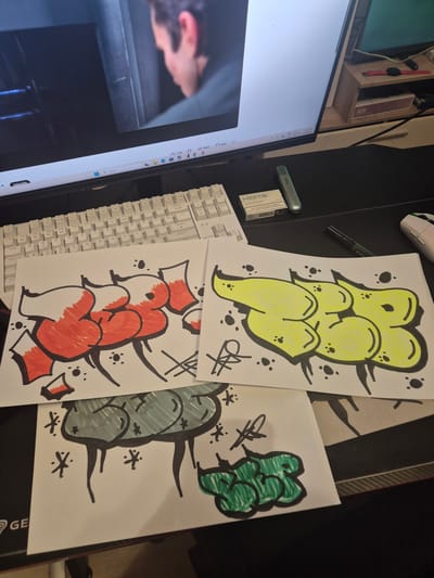 The image shows a desk with a computer monitor displaying a video, a keyboard, and three pieces of paper with graffiti art. The graffiti art consists of bubble letters, one in red and white, one in yellow, and one in green. There are also some smaller drawings on the papers, like stars and scribbles. On the desk, there is also a pack of HEETS and a vape pen. The scene appears to be a home office or gaming setup. There is no visible indication of the location being in Novi han, Bulgaria.