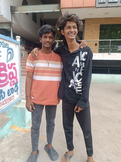 Two young men, likely friends, pose for a photo outdoors in front of a building. The man on the left wears a striped t-shirt with orange, white, and beige horizontal stripes, dark grey jeans, and grey slip-on shoes. He has short black hair and a thin build. The man on the right has long, styled dark hair and wears a black button-down shirt with white text that appears to say "AMERICA" in a repeated pattern, black pants, and flip-flops. He has a ring on his right hand. Both men are smiling, with the man on the right looking off to the side with a wide grin, and the man on the left looking directly at the camera. Their arms are around each other's shoulders.
Behind them, a building with a beige brick facade is visible. A metal railing separates them from the building. To the left of the frame is a blue and white sign with Telugu text and a graphic of a stylized 'D'. Partially visible text on the building includes "CAFE BIRYANI". The ground is paved with concrete, and there are some faded yellow and blue painted markings on the steps leading to the entrance. The lighting suggests it is daytime, likely with bright sunlight, casting some shadows. The overall scene appears to be a casual outdoor setting in a commercial or public area.