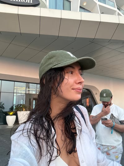 In Ras Al-Khaimah, United Arab Emirates, a woman with wet, dark hair and a white shirt stands in the foreground, facing right. She wears a green baseball cap with a white logo. Behind her, a man wearing a white t-shirt and a green baseball cap with the letter "E" on it, holds a smartphone. He is looking down at the phone. The background features a modern building with large windows and a sign that reads "Hilton". Several potted plants are visible near the entrance. The overall scene suggests an outdoor setting in a hotel area.