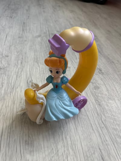 A plastic figurine of a princess, resembling Cinderella, sits on a yellow and purple carriage. The princess wears a blue gown and has orange hair. The carriage has a curved yellow armrest, a purple wheel, and a tan base with white string attached. The figurine is positioned on a light gray wooden floor with visible grain patterns. The scene is devoid of any urban or specific geographical context related to the city of Мариинский, Russia.