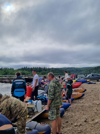 The image depicts a group of people gathered by a body of water, possibly preparing for or concluding a rafting trip. Several inflatable rafts of various colors, including red, blue, and yellow, are present on the shore and in the water. The shore is covered with pebbles. In the background, a dense forest stretches along the opposite bank of the water. The sky is overcast, with some sunlight breaking through the clouds in the upper left corner. A dark-colored car is parked on the right side of the image. People are seen in casual clothing, some of them interacting with the rafts or carrying equipment. One individual is wearing a jacket with "СИБУР" written on the back, a prominent Russian petrochemical company. The scene suggests an outdoor recreational activity taking place in a natural, possibly remote, setting near the city of Vishegorsk, Russia.
