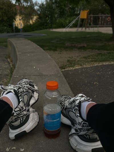 The image shows a person's legs and feet, wearing white socks and chunky black and white sneakers, resting on a concrete curb. To the right of the feet is a partially full plastic bottle of Nestlé Peach Flavor iced tea with an orange cap. In the background, a park or playground area is visible, with green grass, trees, and playground equipment including a yellow structure and a wooden climbing frame. A yellow building is visible behind trees on the left. The setting appears to be an outdoor public space, likely a park or playground, in Roskilde, Denmark, given the context of the request. The lighting suggests it is daytime.