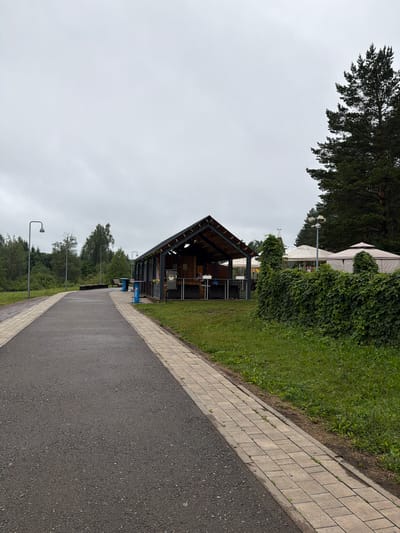 The image depicts an outdoor scene on an overcast day, likely in the afternoon. The setting appears to be a park or a recreational area, possibly a venue for outdoor events.

In the foreground, a paved pathway runs diagonally across the frame, bordered by a brick path on the left. To the left of the brick path, a grassy area slopes upwards, and beyond that, a large open-sided wooden structure with a sloped roof is visible. This structure seems to be a pavilion or a covered seating area, with tables and chairs visible inside. String lights are draped along the roofline of the pavilion, suggesting it might be used for evening events.

Further in the background, lush green trees and dense foliage dominate the landscape. Two large white tents or marquees are partially visible behind the pavilion and amidst the greenery, hinting at a potential event space or temporary structures for an occasion. A streetlamp stands near the tents, and another is visible further down the pathway.

The sky is a uniform gray, indicating cloudy weather. The overall mood is subdued, typical of a gray day. No people are clearly visible in the image. A blue Pepsi branded trash bin is situated near the pavilion. There is no discernible text in the image beyond the Pepsi logo. The location context suggests this scene is in Belebei, Russia.