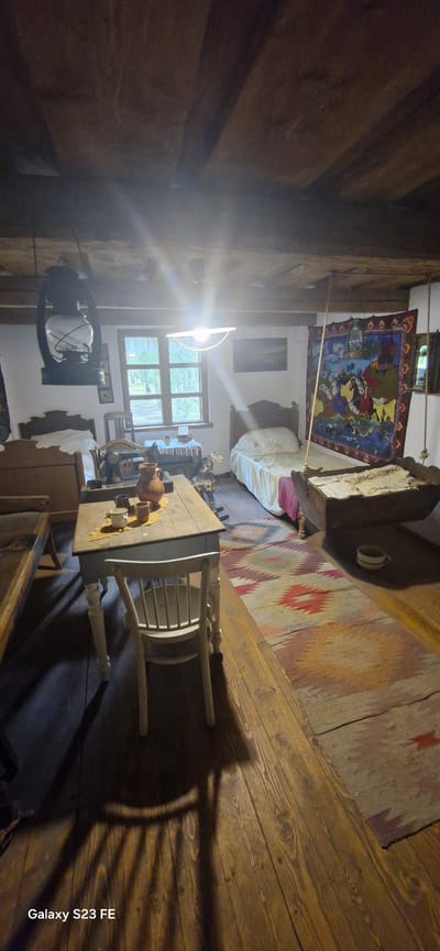 The image depicts the interior of a rustic room, likely a historical dwelling or museum exhibit, situated within the city of Pakruojis, Lithuania. The room is furnished with simple, aged wooden furniture and decorated with traditional textiles.

In the foreground, a wooden table with a light-colored top and turned legs is centrally placed. On the table, there are several earthenware jugs and cups. A white wooden chair with a slatted back is positioned in front of the table, casting a long shadow on the wooden floorboards. The floor itself is made of wide, aged planks, showing wear and tear consistent with age.

To the left of the table, part of a wooden bed frame is visible. Further back, behind the table and slightly to the left, is a large, old-fashioned oil lantern hanging from the ceiling.

In the middle ground, a window with multiple panes looks out onto greenery, suggesting an outdoor scene. A vintage sewing machine sits on a small table or stand in front of the window. A rocking horse, patterned in black and white, stands near the sewing machine. To the right of the window, a bed with white bedding and a pink cloth at its foot is visible.

Dominating the right side of the room, a wooden cradle is suspended from the ceiling by ropes. A colorful, woven tapestry depicting a scene with figures on horseback hangs on the wall behind the cradle. A small, white bowl rests on the floor near the cradle. Another, smaller picture is visible on the wall above the sewing machine.

The ceiling is constructed of rough-hewn wooden beams. The lighting in the room appears to be a combination of natural light from the window and artificial light from a fixture above the window, which casts a bright glare. The overall atmosphere is one of historical preservation and a glimpse into a bygone era.