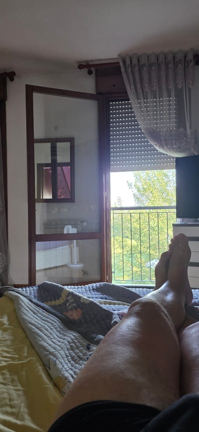 The image is an indoor, eye-level, point-of-view shot from a bed in Montegrotto Terme, Italy. The scene focuses on a person's legs and feet lying on a bed covered with a gray and yellow patterned quilt. In the background, there is a glass door leading to a balcony with lush green trees visible outside. A metal railing is also present on the balcony. To the left of the door, a mirror reflects a staircase and a radiator. The room appears to be a bedroom, possibly in a hotel or residential setting, with sheer curtains hanging by the window.