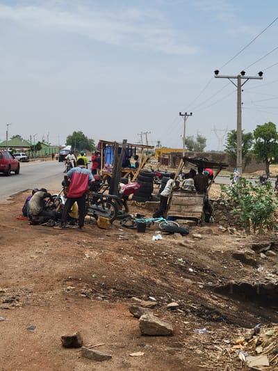 The scene depicts a roadside in Mara, Nigeria. Multiple individuals are gathered around what appears to be a makeshift repair area or small market alongside a paved road. Several motorcycles are present, with at least one individual seemingly working on a motorcycle. The environment is dusty, with dirt and debris along the roadside. There are structures, potentially small shops or stalls, constructed from wood and displaying items for sale. Utility poles and electrical wires are visible overhead. A few vehicles are present on the road, including a red car and white car. Vegetation is sparse, with a few trees in the background.
