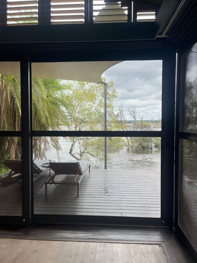 The image depicts a view from inside a building, looking out through a screened window. The exterior features a wooden deck with patio furniture, a table and chair. Beyond the deck is a body of water, likely a lake or river, with trees and vegetation along the shoreline. The sky is overcast and the overall setting suggests an outdoor recreation or residential area. The location is not discernible from the image. The city of Livingstone, Zambia, is not recognizable in the image.
