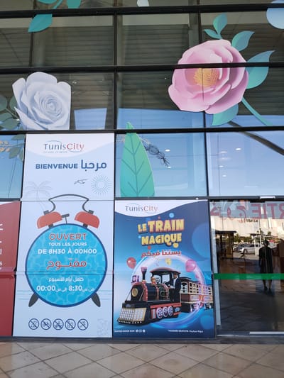 The image shows the exterior of a building with large glass windows, likely a shopping mall or public venue. The windows are decorated with large, stylized paper flowers in white and pink, and green leaf cutouts.

On the left side, there is a large poster with the text "TunisCity" and "BIENVENUE" in large blue letters, accompanied by Arabic script. Below this, there is an illustration of an alarm clock with the text "OUVERT TOUS LES JOURS DE 8H30 À 00H00" and Arabic text stating "مفتوح كامل أيام الأسبوع من س 8:30 إلى س 00:00". At the bottom of this poster, there are icons depicting prohibited activities: a dog, a bicycle, a stroller, a bottle, and a person smoking.

In the center, there is a poster featuring a colorful illustration of a toy train with the text "LE TRAIN MAGIQUE" in yellow letters and Arabic text below it. Balloons are depicted around the train. At the bottom of this poster, it says "SUIVEZ-NOUS SUR:" followed by social media icons and "TOURNÉE GRATUITE / جولة مجانية".

To the right of the posters, there is a glass entrance with a green band across it. Reflections in the glass show cars and people inside a brightly lit space. The ground in front of the building is paved with square tiles. The overall impression is of an advertisement for a shopping center in Tunis City, Tunisia, highlighting its hours of operation and a children's train attraction.
