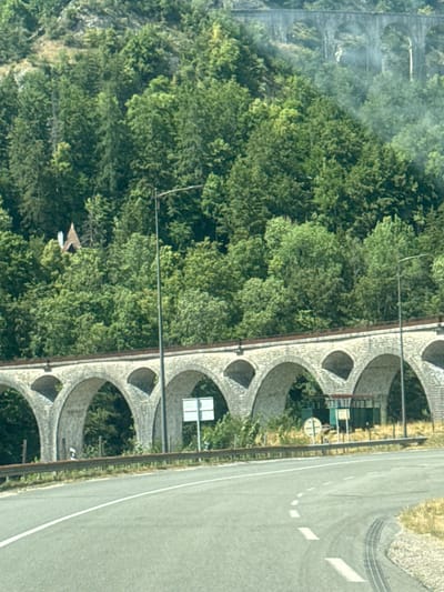 The image shows a road in the foreground with a white line marking a lane. In the background, a stone viaduct with multiple arches runs across the landscape, spanning a wooded area. The viaduct is built on a slope, and trees grow densely on the hillsides. A small building with a distinctive roof is visible nestled amongst the trees on the left side of the viaduct. A street lamp is positioned near the viaduct, and various road signs are present. The overall scene depicts a rural or mountainous area with a historical structure. The location is identified as the city of Hauts de Bienne, France.