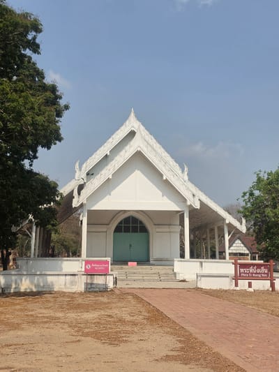 The image shows Phra Thinang Yen, a white building with a light green door in Phra Nakhon Si Ayutthaya City Municipality, Thailand. The building has a triangular roof with ornate details. There are trees on the left, and a brick path to the right. Signs indicate that entry is restricted. The sky is blue with some clouds.