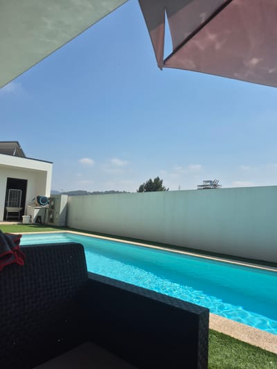 The image depicts a modern outdoor swimming pool area, likely located in Felgueiras, Portugal, on a clear day. In the foreground, a dark woven armchair is partially visible, with a red and gray cloth draped over its arm. The swimming pool, with its bright turquoise water, occupies a significant portion of the mid-ground, reflecting the sunlight and showing gentle ripples. The pool is surrounded by artificial green turf.
To the left of the pool, a white modern building is partially visible, featuring a dark doorway and a large white air conditioning unit or cooler. Adjacent to this, two outdoor air conditioning units are mounted on the wall.
A tall, pale green wall forms a backdrop across the mid to background of the scene, separating the pool area from the surrounding landscape. On top of this wall, in the distance, a structure resembling lounge chairs on a metal frame is visible. Further in the background, a line of trees and hills can be seen against the clear blue sky, with a few wispy clouds scattered across it. An umbrella, colored in shades of pink and purple, is angled down from the top right corner of the frame, casting a shadow on the sky.
