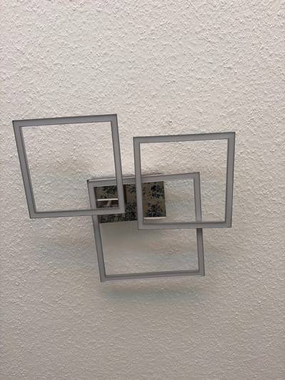 This media file shows a modern wall lamp mounted on a textured white wall. The lamp consists of three square frames of varying sizes, arranged in an overlapping, geometric design. The frames themselves appear to be made of a light-colored material and are illuminated from within, casting a soft, diffused light. A central metallic element, possibly the mounting bracket or a decorative piece, is visible behind the overlapping frames. This central piece has a marbled or speckled pattern, featuring dark and light grey tones with hints of blue. The texture of the wall is rough and uneven, with small raised bumps and crevices, creating shadows and highlighting the depth of the surface. The lighting in the scene is gentle, suggesting it might be daytime with diffused light, or the lamp itself is the primary light source in a dimly lit room. There is no visible text in the image. The overall impression is one of contemporary design and subtle illumination in a domestic setting.