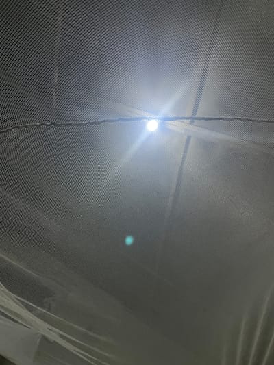 The image shows a close-up view of a white mesh fabric, likely a mosquito net, with a bright light source directly above and slightly to the left, creating a lens flare. The fabric has a regular pattern of small holes. There is a dark line running horizontally across the middle of the frame, possibly a seam or a tie. A faint green blur is visible in the lower middle portion of the image. The overall impression is of looking through a transparent barrier at a light source. There are no people, distinct objects, or specific environmental details visible that would suggest a location in Omu Aran, Nigeria.