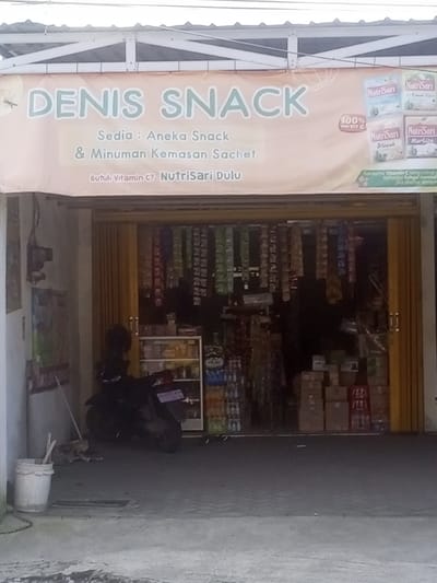 The image shows the exterior of a small shop named "Denis Snack," located in Kecamatan Waru, Indonesia at coordinates -7.35 latitude and 112.764 longitude. The shop has a yellow awning with the name of the shop and the words "Sedia: Aneka Snack & Minuman Kemasan Sachet" and "Butuh Vitamin C? Nutrisari Dulu". The shop window is filled with various packaged goods and there are several boxes of goods stacked on the right side of the shop. A black motorcycle is parked in front of the shop with a white bucket nearby. The shop is situated on a concrete floor and there is a grey wall on the left side of the image. The awning is partially obscured by an overhang above the shop and the weather appears overcast.