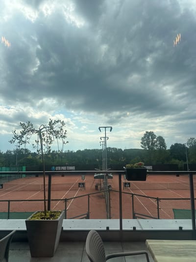 The image displays an elevated view of multiple clay tennis courts located in Giebułtów, Poland, under a predominantly overcast sky. The immediate foreground shows a light-colored patio surface, a glass and dark metal railing, a tall, slender plant in a dark rectangular planter on the left, and a dark hanging planter containing red flowers on the right. Parts of a grey outdoor chair and a light-colored table are visible at the bottom edges.
Beyond the railing, at least two reddish-brown clay tennis courts are present, each equipped with white boundary lines and nets. A black banner with white text "UTR PRO TENNIS" is stretched along the rear fence of the nearest court. Several smaller black and white signs, also featuring "UTR PRO TENNIS" branding, are positioned around the courts. Tall, multi-fixture light poles stand behind the courts. Green mesh fencing separates the playing areas. The background consists of a dense line of green trees. The sky, occupying the upper two-thirds of the frame, is heavily clouded with shades of grey, though some brighter areas of sky are visible towards the horizon. Reflections are present on the glass barrier in the upper portion of the image. No individuals are depicted on the courts or in the surrounding environment.