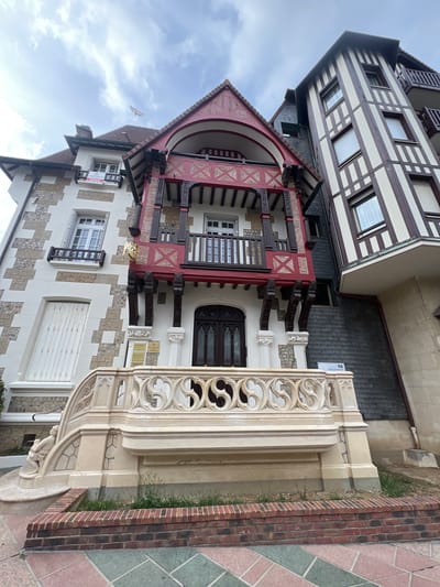 This is a medium shot, low-angle, eye-level, full-color, outdoor, daylight photograph of the exterior of a building in Deauville, France. The building appears to be a mix of architectural styles, with a portion featuring a half-timbered design and another with a lighter-colored stone and stucco facade. A prominent, ornate balcony with intricate stone carvings and a decorative railing is in the foreground, leading up to the entrance of the building. The balcony is supported by decorative columns and appears to be made of stone or a similar material. Above the balcony, a vibrant red and dark wood balcony juts out from the main structure, adorned with decorative woodwork. The building on the right displays a classic half-timbered facade with dark brown beams against a white background. The sky is overcast with some clouds visible. A single bird is captured in flight in the upper left quadrant of the image. A "VENDU" (SOLD) sign is visible on a window of the building on the left, suggesting a real estate transaction. The ground in front of the building is paved with colored geometric tiles, and a low brick wall encloses a small patch of grass. The building is located on a street in Deauville, France, a resort town known for its architecture and association with horse racing and film festivals.