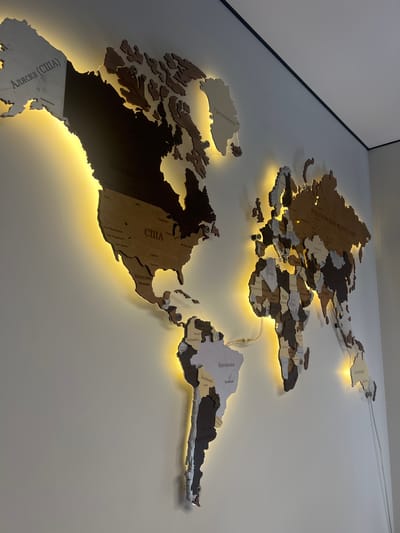 This is a well-lit, close-up, eye-level shot of a wooden world map art piece mounted on a white wall. The map features various continents and countries intricately cut and assembled, with some sections being lighter wood and others darker. The pieces are outlined with a warm yellow LED light that casts a glow around the edges. Several countries and cities are labeled in Cyrillic script. Prominently visible labels include "Аляска (США)" (Alaska, USA), "Канада" (Canada), "США" (USA), "Мексика" (Mexico), "Российская Федерация" (Russian Federation), "Австралия" (Australia), "Бразилия" (Brazil), and "Аргентина" (Argentina). Some of the cities labeled within the countries include "Сан-Франциско" (San Francisco), "Лос-Анджелес" (Los Angeles), "Сан-Диего" (San Diego), "Хьюстон" (Houston), "Даллас" (Dallas), "Москва" (Moscow), "Екатеринбург" (Yekaterinburg), "Кишинев" (Chisinau), "Анкара" (Ankara), "Каир" (Cairo), "Нагер" (N'Djamena), "Мали" (Mali), "Нджамена" (N'Djamena), "Судан" (Sudan), "Аддис-Абеба" (Addis Ababa), "Богота" (Bogota), "Колумбия" (Colombia), and "Лима" (Lima). There are visible wires connected to the LED lights, one of which snakes down the wall to the right. The wall appears clean and the room is dimly lit except for the illumination from the map's LEDs. The top right corner of the image shows a thin black trim line where the wall meets the ceiling. There are no people or animals present.