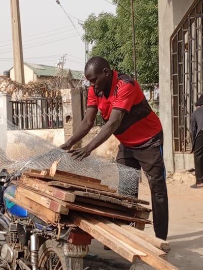 A daylight scene in Kwachiri, Nigeria, depicts an adult male positioned beside a heavily loaded motorcycle on an unpaved surface. The man, of African descent, wears a red short-sleeved t-shirt with black horizontal stripes and dark-colored athletic pants with a light blue stripe. He is leaning forward, with both hands resting on a large roll of silver-colored wire mesh that is part of the cargo. Beneath the wire mesh, a stack of various wooden planks is secured to the rear of a blue and silver motorcycle, partially visible on the left.
In the background, multiple structures are visible under a clear sky. To the immediate right of the primary subject, a light-colored building with a dark metal security gate is present. Further back, a building with a green roof is seen, along with additional residential or commercial structures and a utility pole on the far left. Green trees with dense foliage are prominent in the mid-ground and background. A second individual, wearing dark pants and shoes, is partially visible standing on the far right edge of the frame.