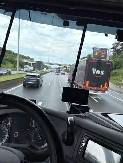 This media file presents a driver's view from inside a vehicle, likely a truck, looking forward onto a multi-lane highway near Kortrijk, Belgium. The perspective is from the driver's seat, showing part of the dashboard, steering wheel, and windshield.
In the immediate foreground, the right side of the dashboard is visible, featuring a digital display unit, possibly an infotainment system, and a mounted smartphone or navigation device (labeled "Garmin"). To the left, through the windshield, traffic flows on a busy highway under an overcast sky.
The highway is populated with numerous vehicles, primarily cars and large trucks. Directly ahead of the viewer's vehicle is a black semi-trailer truck. The back of this truck clearly displays the branding "VOS Logistics". Further down the road, more vehicles are visible in multiple lanes, some appearing to be in slower-moving traffic or potentially an exit lane. An overpass crosses the highway in the mid-distance.
A prominent feature on the left side of the highway is a large digital variable message sign (VMS). This sign displays a red circle with "70" inside, indicating a speed limit of 70 km/h, with "2 km" below it, signifying the duration of this limit. To the left of the speed limit, a large yellow "1" is displayed, which might relate to lane information or an incident. The road surface appears damp in places, suggesting recent rain or general wet conditions. The time of day is daylight, characterized by the natural light despite the cloudy weather. The vehicle's dashboard instrumentation is partially visible, showing gauges and digital readouts.